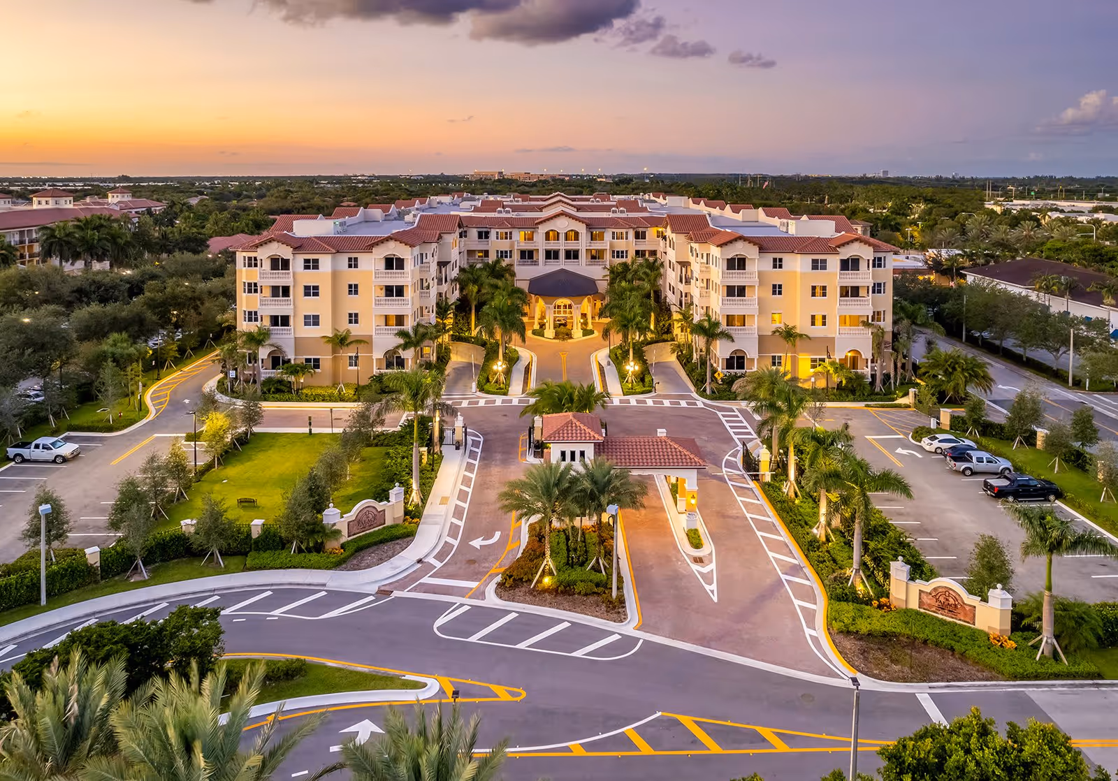 Aerial view of The Palace at Weston senior living facility at sunset, showing a large, multi-story building with a red-tiled roof, palm trees lining the entrance driveway, and surrounding parking areas and greenery.