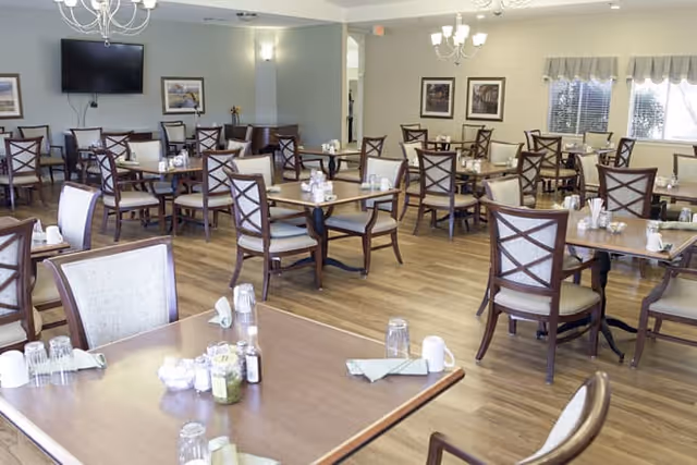 A spacious dining room with multiple wooden tables and chairs arranged neatly. Each table is set with glasses, napkins, and condiments. The room has light-colored walls, framed pictures, a wall-mounted TV, and windows with valances allowing natural light to enter.