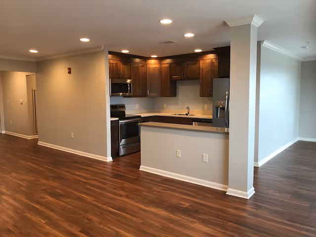 Open-plan interior showing a modern kitchen with wooden cabinets, stainless steel appliances, and a breakfast bar opening to a hardwood-floored living area.