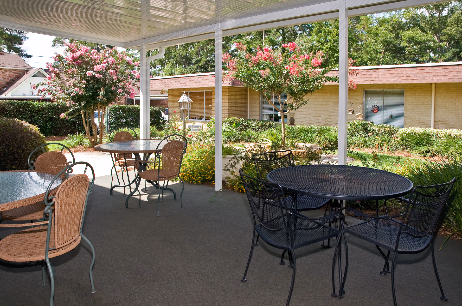 Covered outdoor patio with round tables and chairs overlooking a landscaped courtyard and a single-story building.
