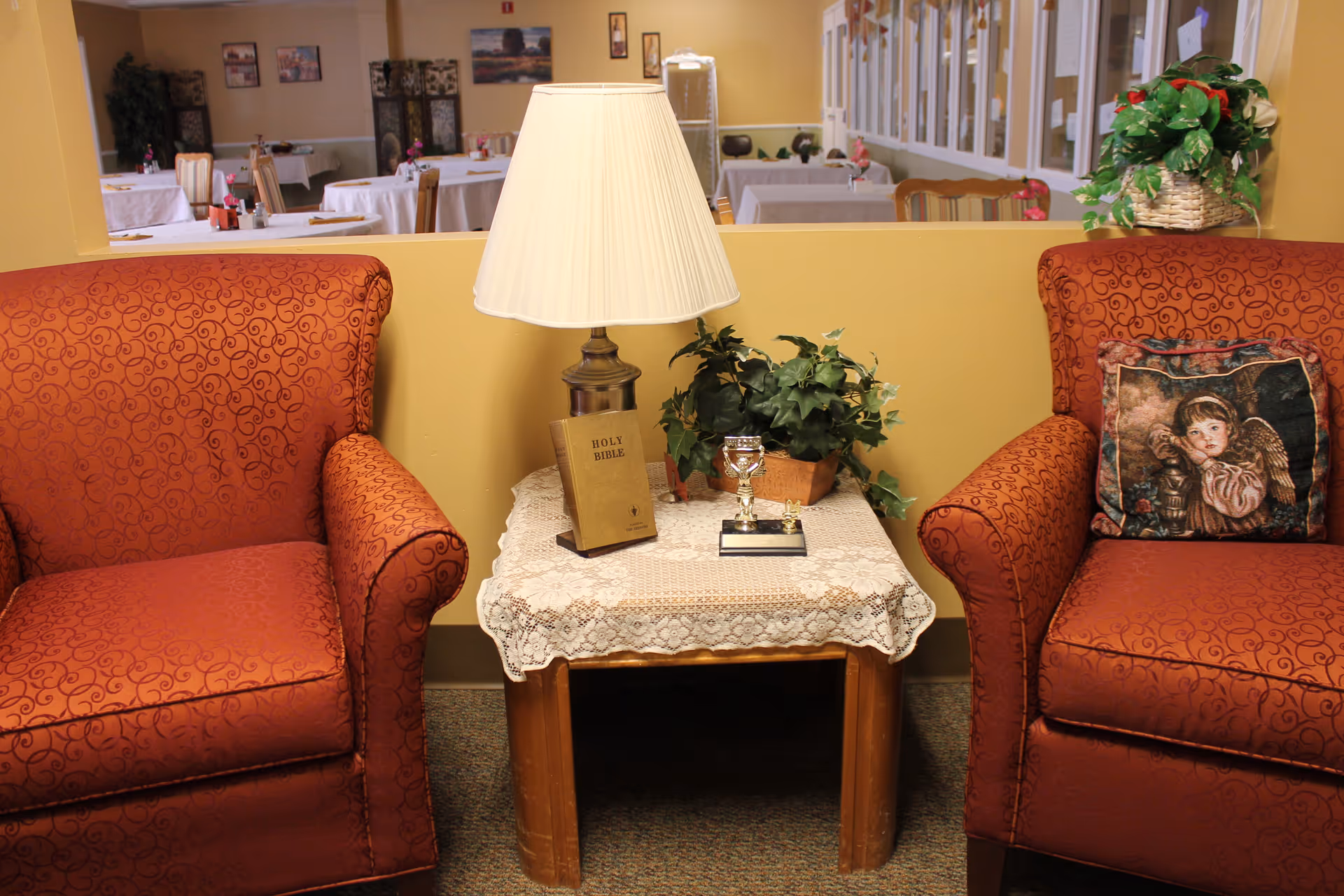 Two red patterned armchairs with a small wooden table between them. The table is covered with a lace cloth and holds a lamp, a Holy Bible, a small trophy, and a potted plant. Behind the chairs is a window looking into a dining area with tables and chairs.