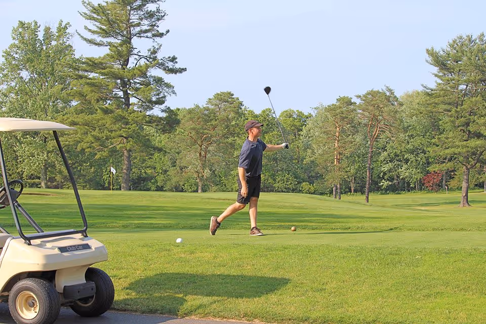 A man playing golf on a green golf course with trees in the background and a golf cart parked nearby.