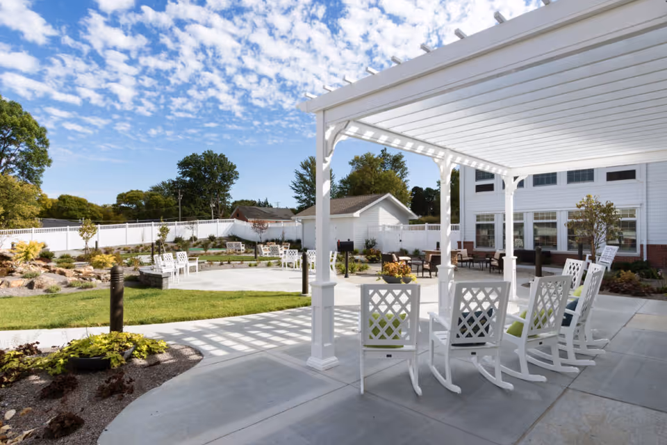 Outdoor patio area at CountryHouse Residence featuring white rocking chairs under a white pergola, a concrete walkway, landscaped garden with small trees and shrubs, and a white fence enclosing the space under a partly cloudy blue sky.