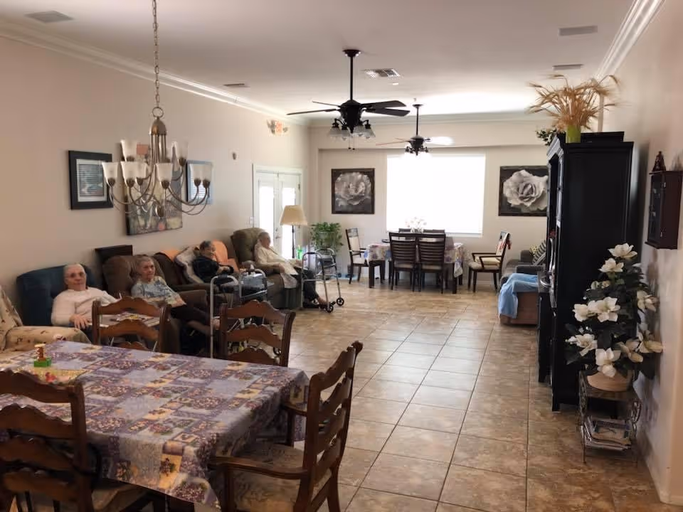 A spacious living room in an assisted living facility with several elderly residents seated on sofas and chairs along the left wall. The room features tiled flooring, ceiling fans, a dining table with chairs covered by a patterned tablecloth in the foreground, and another dining area with chairs near a large window at the back. The walls are decorated with framed floral artwork, and there are plants and flowers placed around the room.