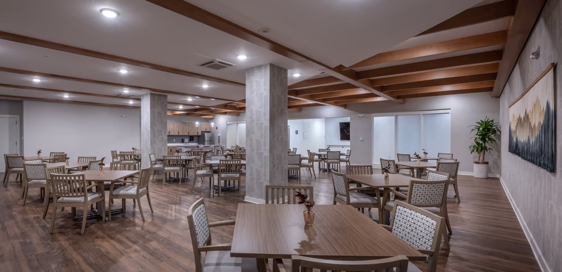 A spacious dining room with multiple wooden tables and chairs arranged neatly. The room features wooden flooring, exposed wooden beams on the ceiling, and soft overhead lighting. There are two large pillars in the center, a kitchen area with cabinets and appliances in the background, a large wall hanging artwork, and a potted plant in the corner.
