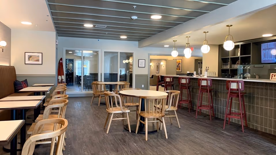 Bright communal dining area with round and rectangular tables, wooden chairs, and a bar with red stools and pendant lights.