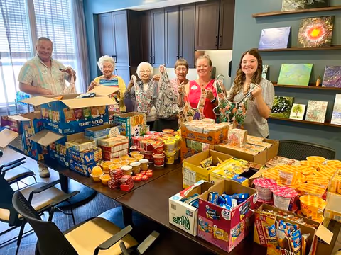 Six people standing behind a table filled with various packaged food items and snacks in a room with dark cabinets and shelves displaying artwork. They are holding reusable bags and smiling at the camera.