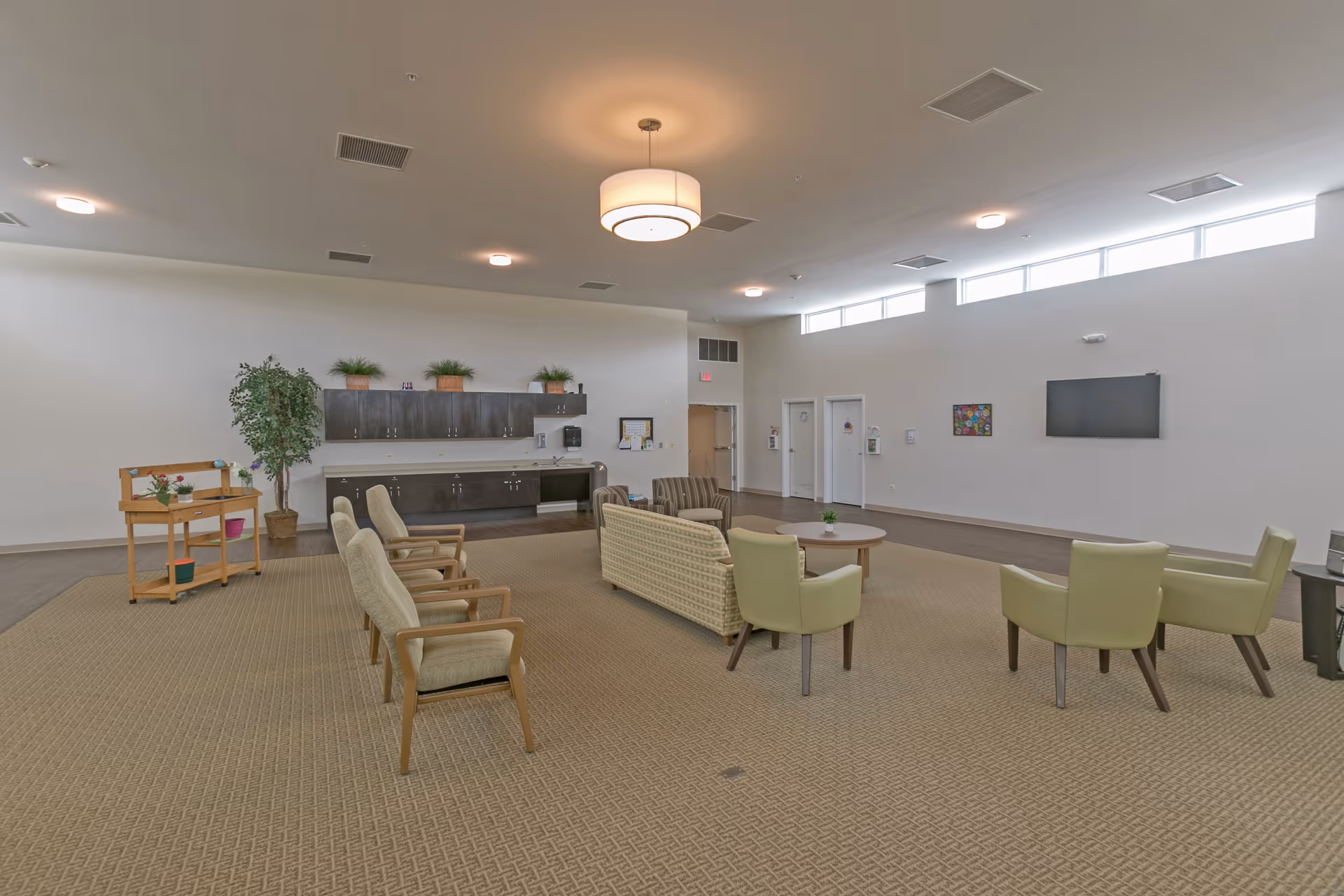 A spacious common area with beige carpet and neutral-colored walls. The room features a seating arrangement with a sofa, armchairs, and a coffee table in the center. On one side, there is a wooden cart with plants and decorative items, and a large potted plant next to a wall with dark cabinets and a countertop. The room is well-lit with ceiling lights and natural light coming through high windows. A flat-screen TV is mounted on the wall, and there are doors leading to other rooms.