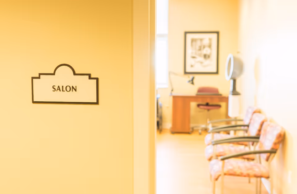 View of a salon room in a senior living facility with a sign labeled 'SALON' on a yellow wall. Inside the room, there are three patterned chairs lined up against the right wall, a wooden desk with a red chair, a desk lamp, and a hair dryer hood. A framed black and white picture hangs on the back wall.