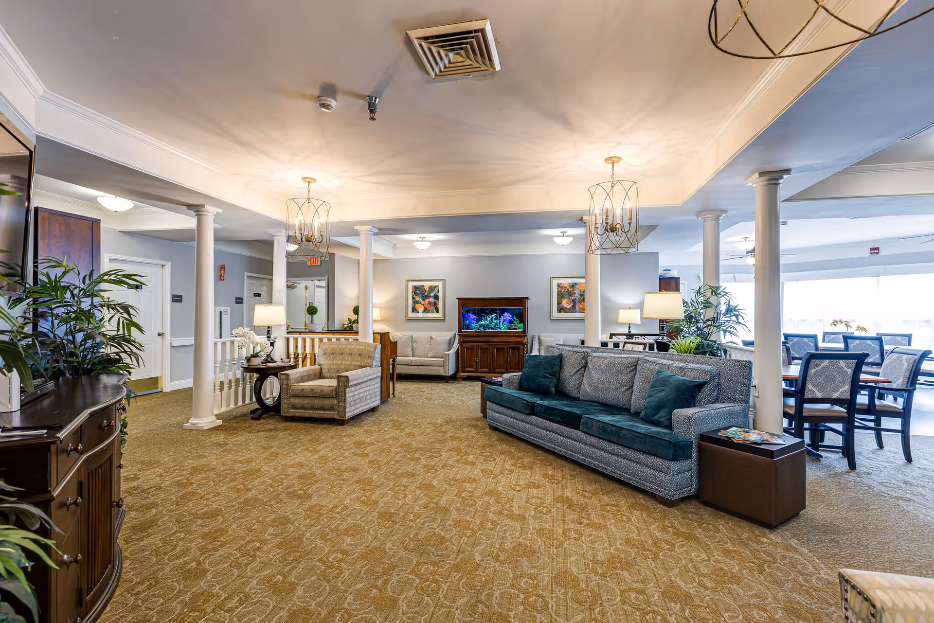 A spacious senior living facility common area with beige carpet, multiple seating options including a gray sofa with teal cushions, armchairs, and a dining table with chairs near large windows. The room features white columns, decorative ceiling lights, plants, and a wooden cabinet with a fish tank and framed artwork on the wall behind it.