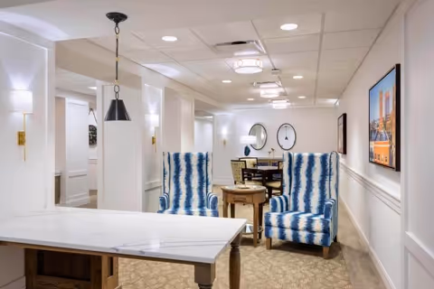 A well-lit interior room featuring a white marble table in the foreground, two blue and white patterned armchairs with a small wooden side table between them, and a dining table with chairs in the background. The walls are white with decorative molding, and there are framed pictures and round mirrors on the walls. Ceiling lights and wall sconces provide illumination.