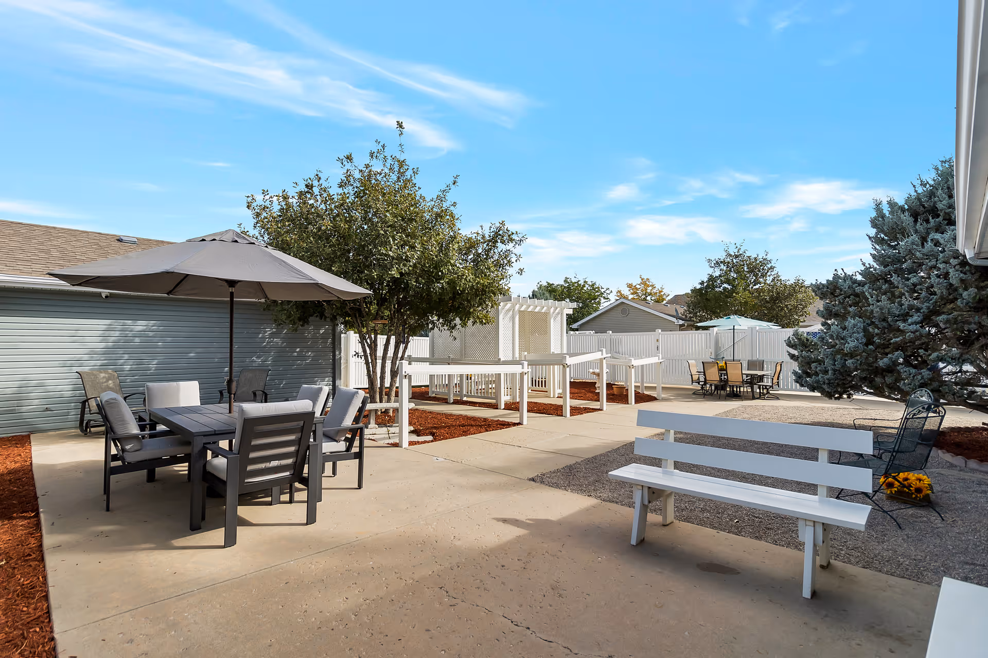 Outdoor patio area with seating arrangements including a table with an umbrella and chairs, a white bench, and another table with chairs in the background. There are trees, a white fence, and a clear blue sky.