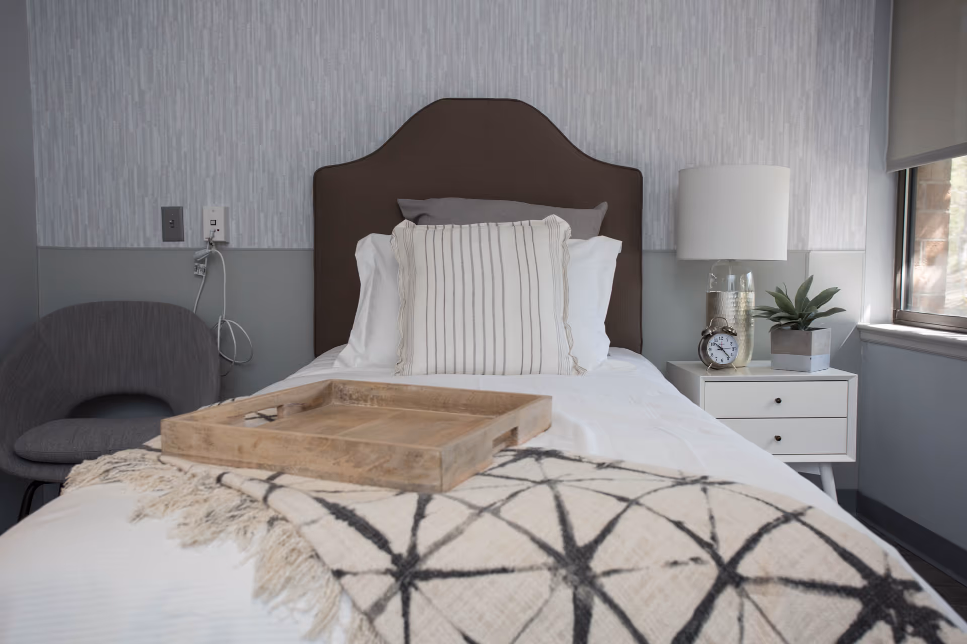 A neatly made single bed with a brown upholstered headboard, white and gray pillows, and a beige throw blanket with a black geometric pattern. A wooden tray is placed on the bed. To the right of the bed is a white nightstand with a table lamp, a small clock, and a potted plant. A gray chair is positioned to the left of the bed, and a window with a roller shade is on the right wall.