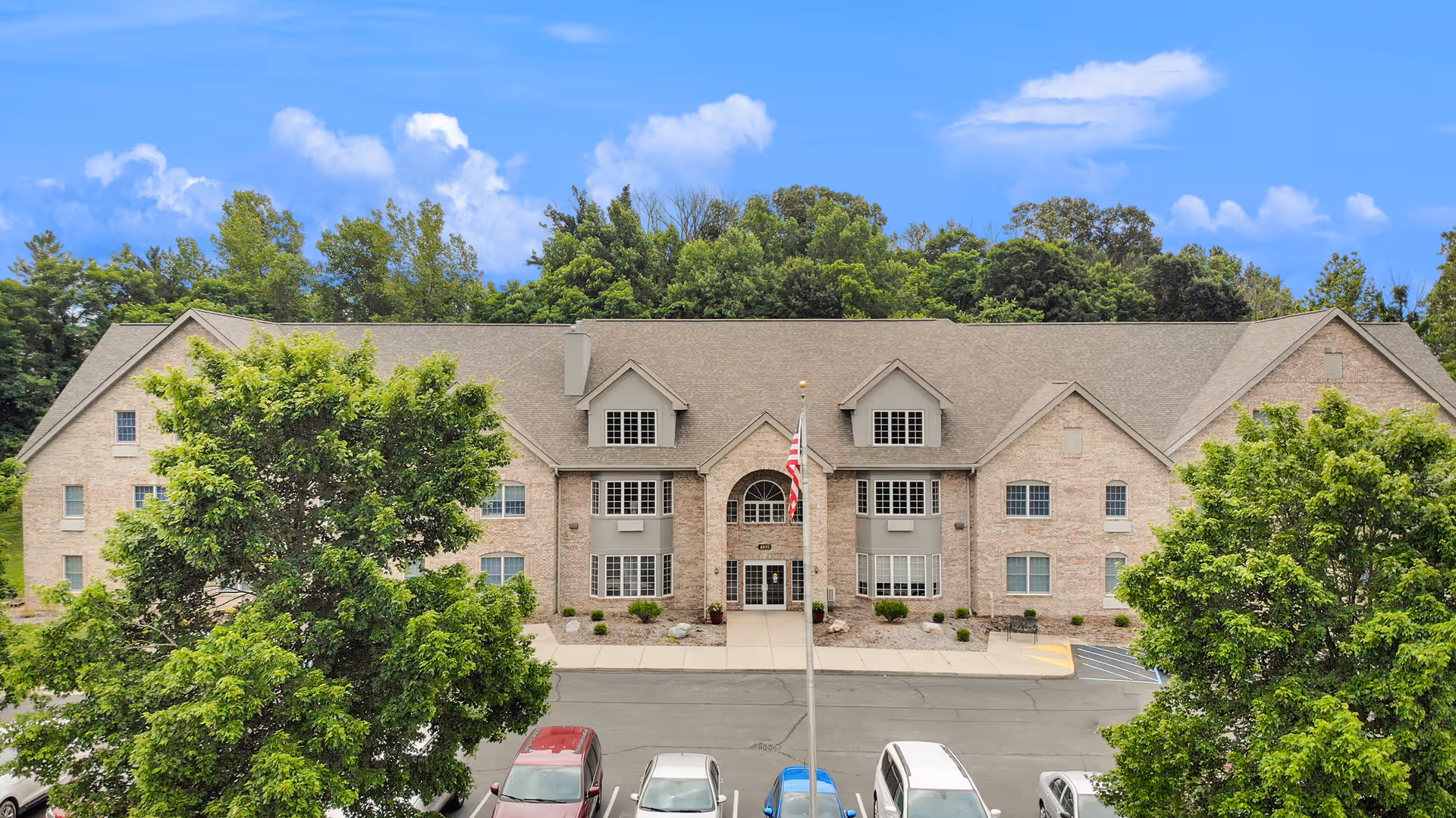 Front exterior view of a two-story brick building with multiple windows and a central entrance. There is a flagpole with an American flag in front of the building, surrounded by trees and a parking lot with several cars parked.