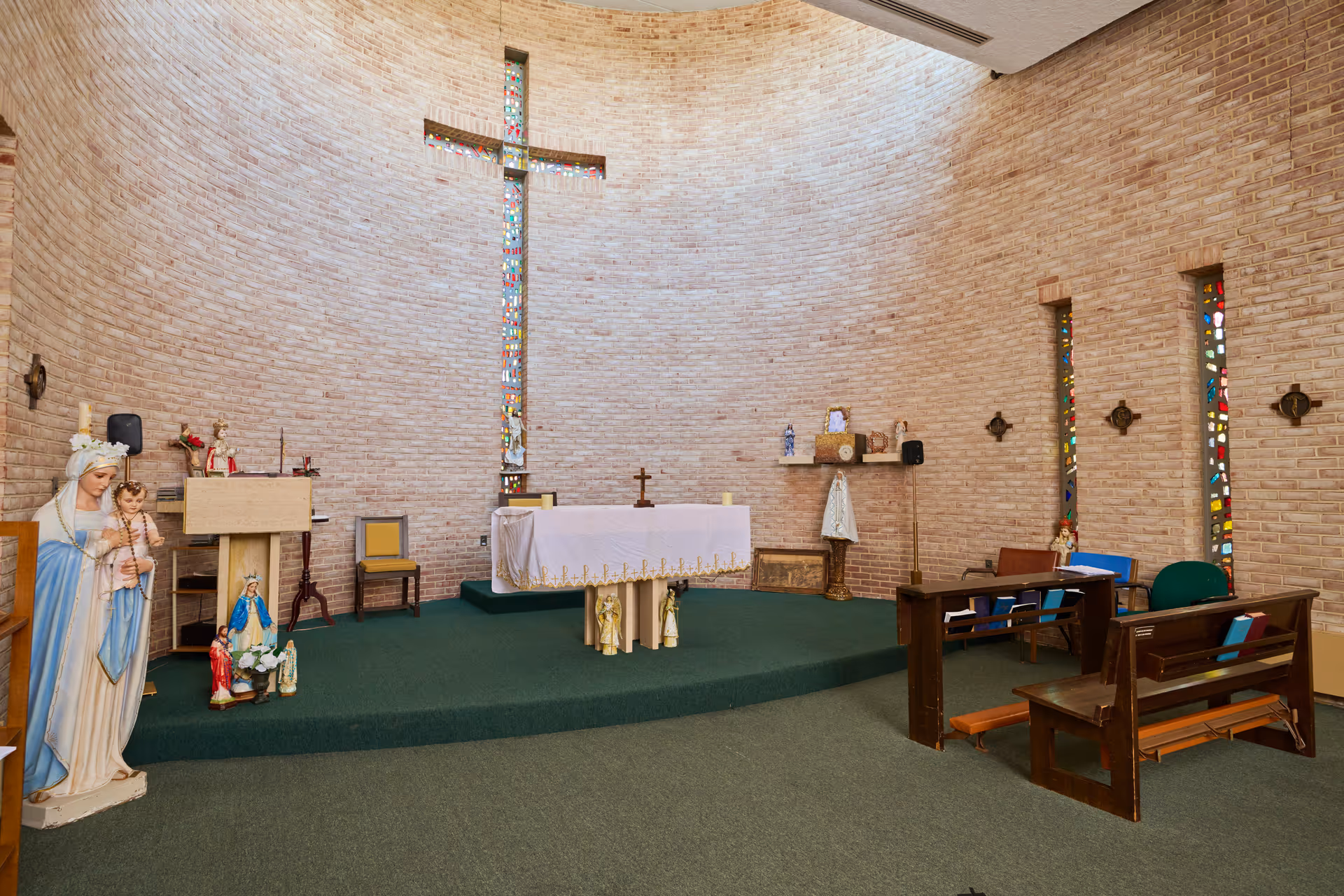 Interior of a small chapel with brick walls and a green carpeted floor. There is a raised platform with an altar covered by a white cloth with gold trim. Behind the altar is a tall cross-shaped stained glass window. To the left, there is a statue of the Virgin Mary holding baby Jesus and other religious statues. Wooden pews are arranged on the right side of the chapel with hymnals and a green chair nearby.