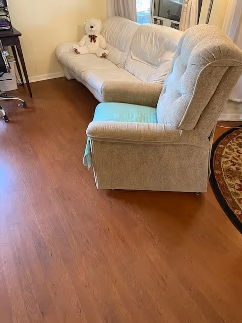 A cozy living room area with a beige armchair in the foreground and a white couch with a white teddy bear sitting on it against a light yellow wall. There is a wooden floor and a round patterned rug partially visible on the right side. A desk with a chair is seen on the left side near the wall.