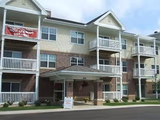 Front exterior of a three-story senior apartment building with balconies, a covered entrance, and a 'Now Leasing' banner.