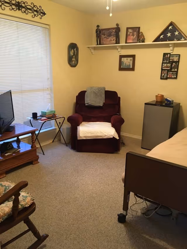 A cozy senior living room with a maroon armchair covered with a white towel and a gray blanket draped over the back. A small refrigerator stands next to the chair, and a bed with wheels is partially visible on the right. A wooden rocking chair with a floral cushion is in the foreground. A TV sits on a wooden stand near a window with closed blinds. The walls are decorated with family photos, a folded American flag in a triangular display case, and small figurines on a white shelf.