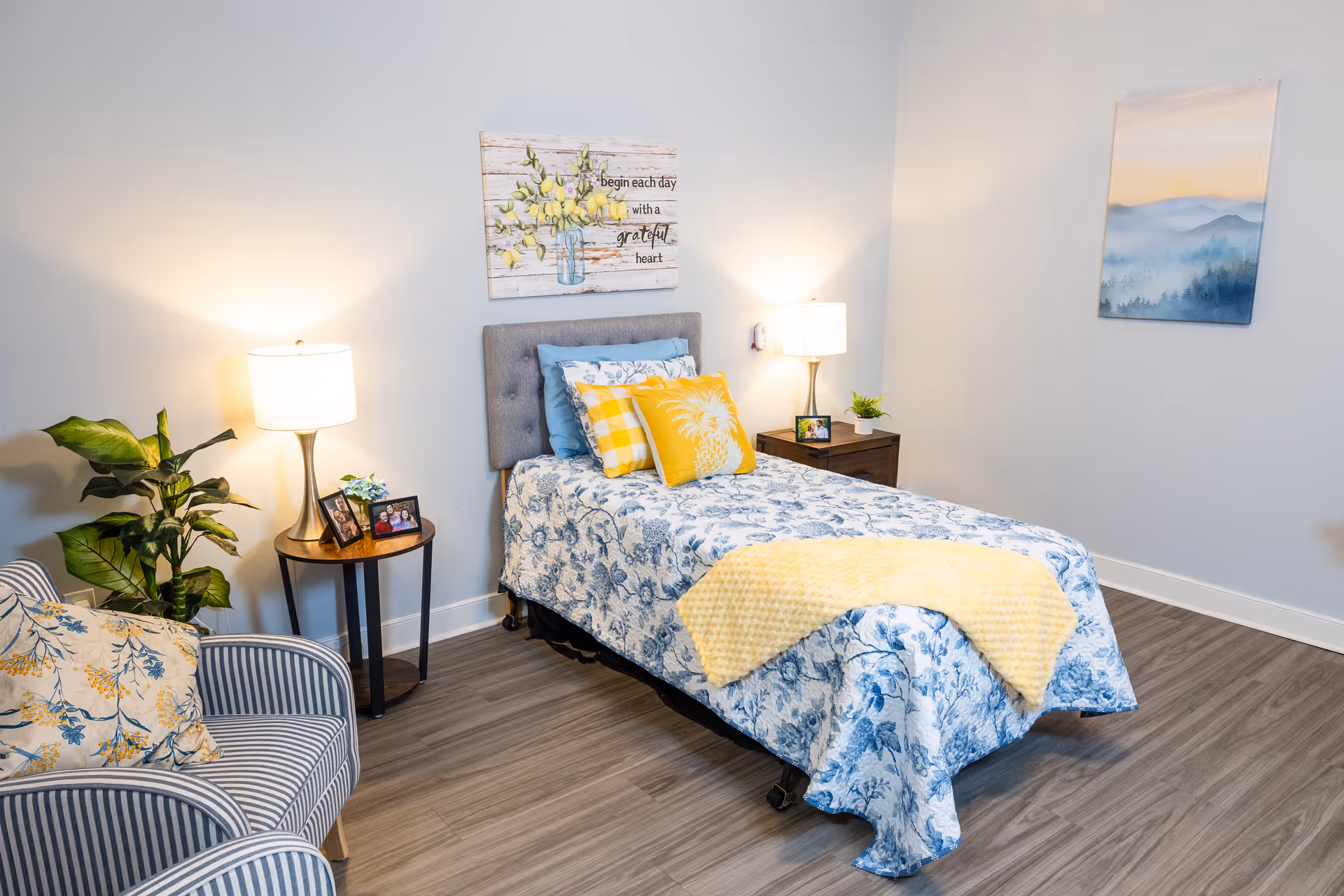 Well-lit single bedroom with a twin bed in blue-and-white floral bedding, yellow accent pillows and throw, bedside tables with lamps, and a striped armchair.