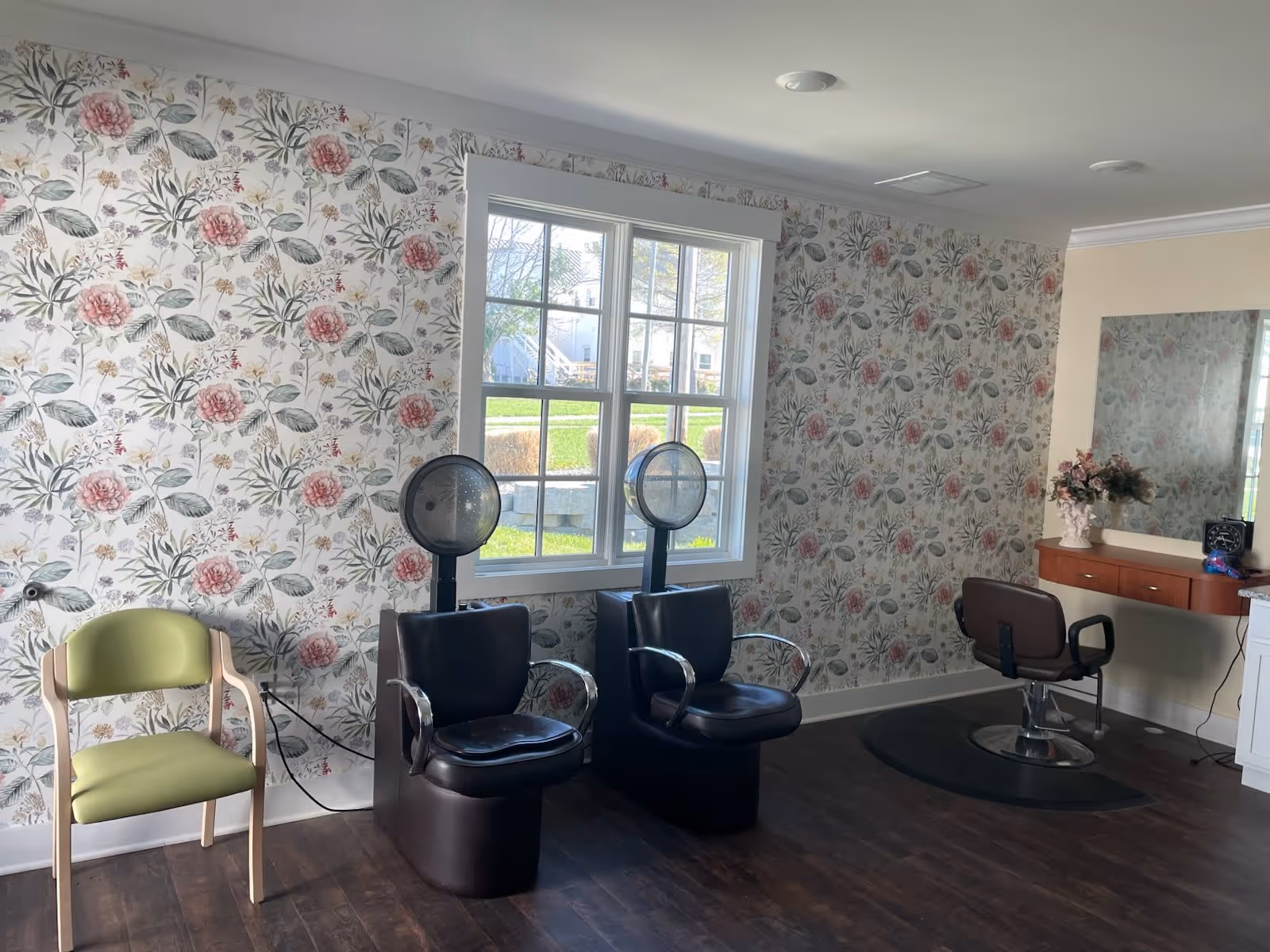 Interior room with floral wallpaper featuring pink and green flowers, two black salon hair dryer chairs, a green cushioned chair, a large window showing an outdoor view, and a vanity area with a mirror, a brown salon chair, and a vase with flowers.