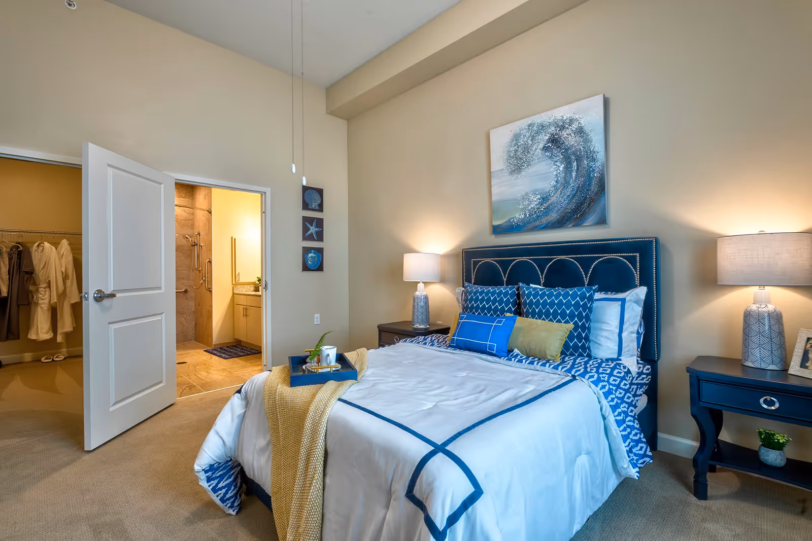 A neatly made bed with blue and white bedding and multiple decorative pillows in a bedroom. Two bedside tables with lamps flank the bed. A painting of a wave hangs above the headboard. An open door reveals a walk-in closet with hanging robes and a bathroom with a shower and vanity.