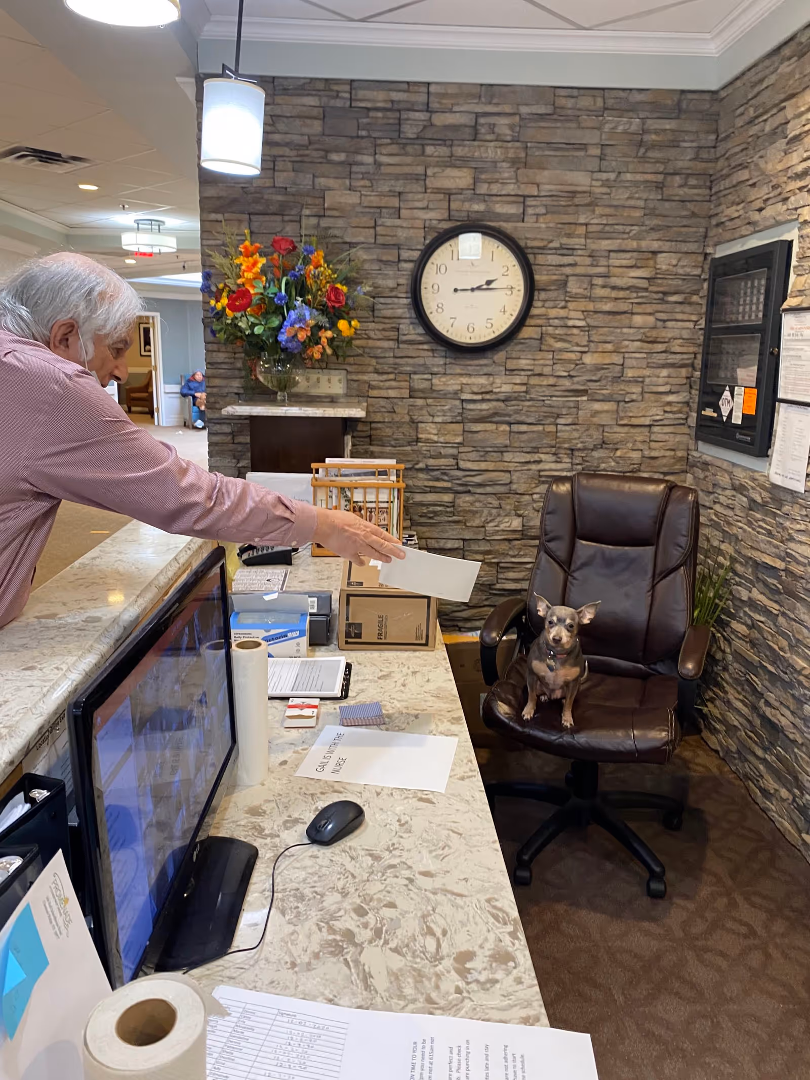An elderly man wearing a mask is reaching across a reception desk to hand a piece of paper to a small dog sitting on a brown leather office chair. The background features a stone wall with a round clock showing the time as 2:15, a colorful flower arrangement, and a bulletin board with papers pinned to it.