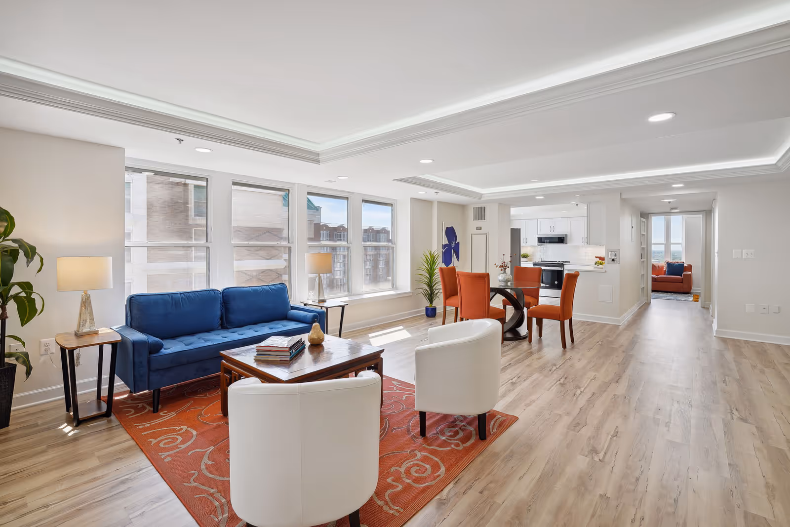 Bright and spacious living area with large windows letting in natural light. The room features a blue sofa, two white armchairs, a wooden coffee table on an orange patterned rug, and a dining area with a round glass table and four orange chairs. The background shows a modern kitchen with white cabinets and stainless steel appliances, and a hallway leading to another room with an orange sofa.