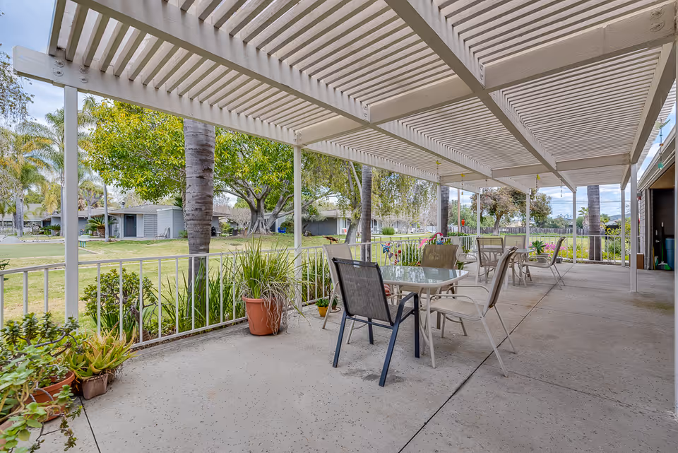 Covered outdoor patio area with several glass-top tables and chairs arranged for seating. The patio overlooks a grassy area with trees and shrubs, and there are potted plants placed along the edge of the patio. The sky is partly cloudy.