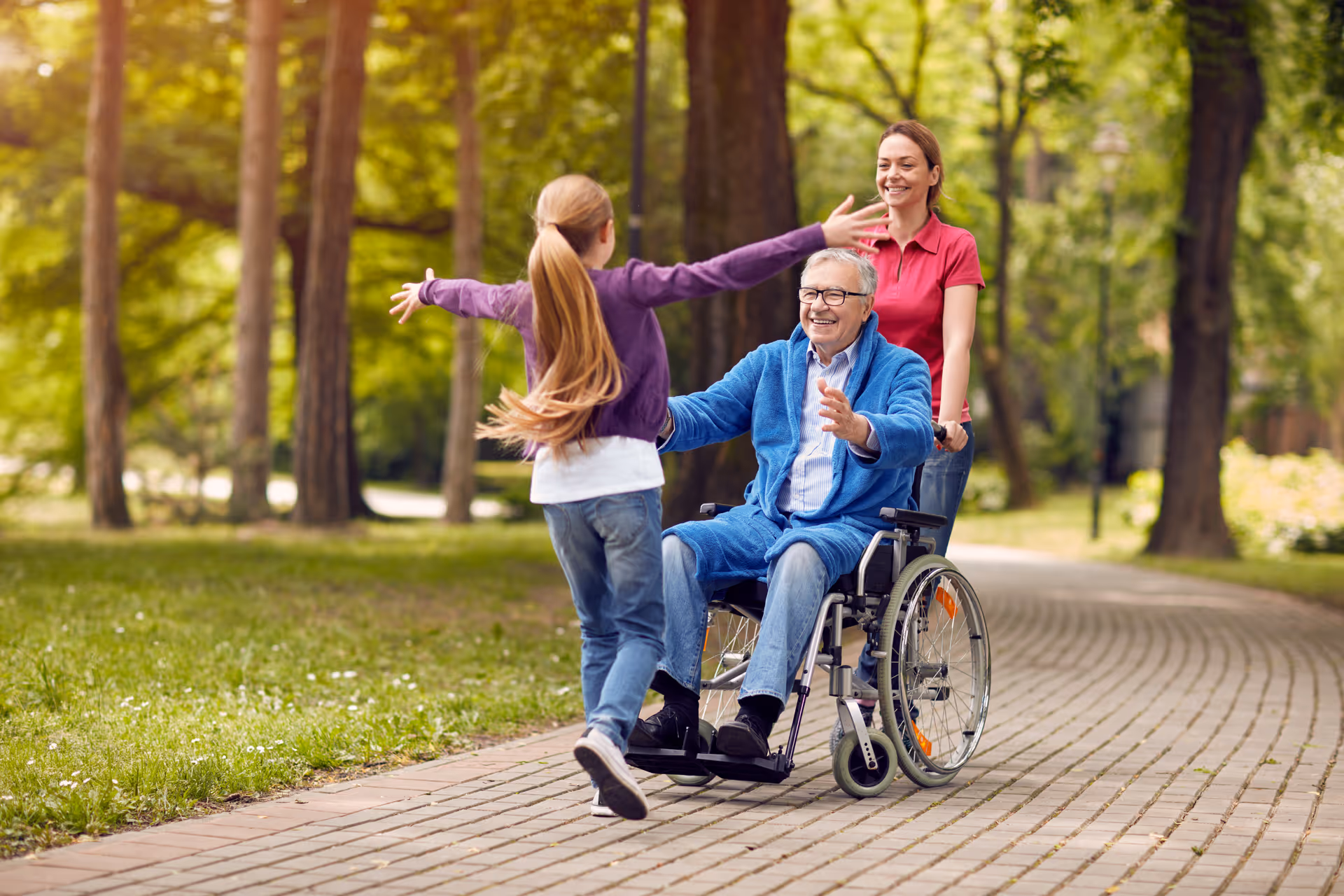 An elderly man in a wheelchair is being pushed by a woman along a paved path in a park. A young girl with long hair is running towards the man with open arms, and all three are smiling. The background shows green trees and grass in a sunny outdoor setting.