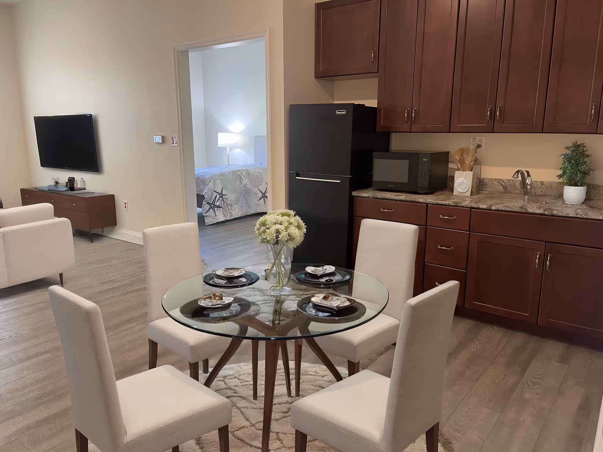 A modern assisted living apartment interior featuring a glass dining table with four beige upholstered chairs set with plates and napkins. The kitchen area has dark wood cabinets, a black refrigerator, a microwave, a sink, and a small potted plant on the counter. In the background, a living area with a wall-mounted TV and a beige armchair is visible, along with a bedroom that has a bed with a starfish-patterned bedspread and a lit bedside lamp.