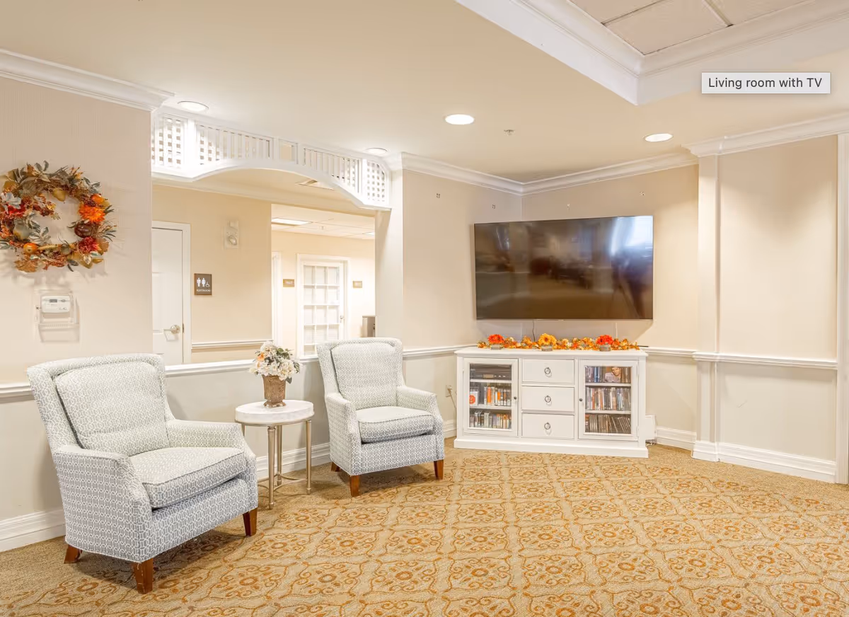 A cozy living room area with two patterned armchairs separated by a small round table with a flower vase. A large flat-screen TV is mounted on the wall above a white cabinet decorated with autumn-themed garland. The room has beige walls, a patterned carpet, and recessed ceiling lights. A decorative wreath hangs on the wall near a doorway labeled as restrooms.