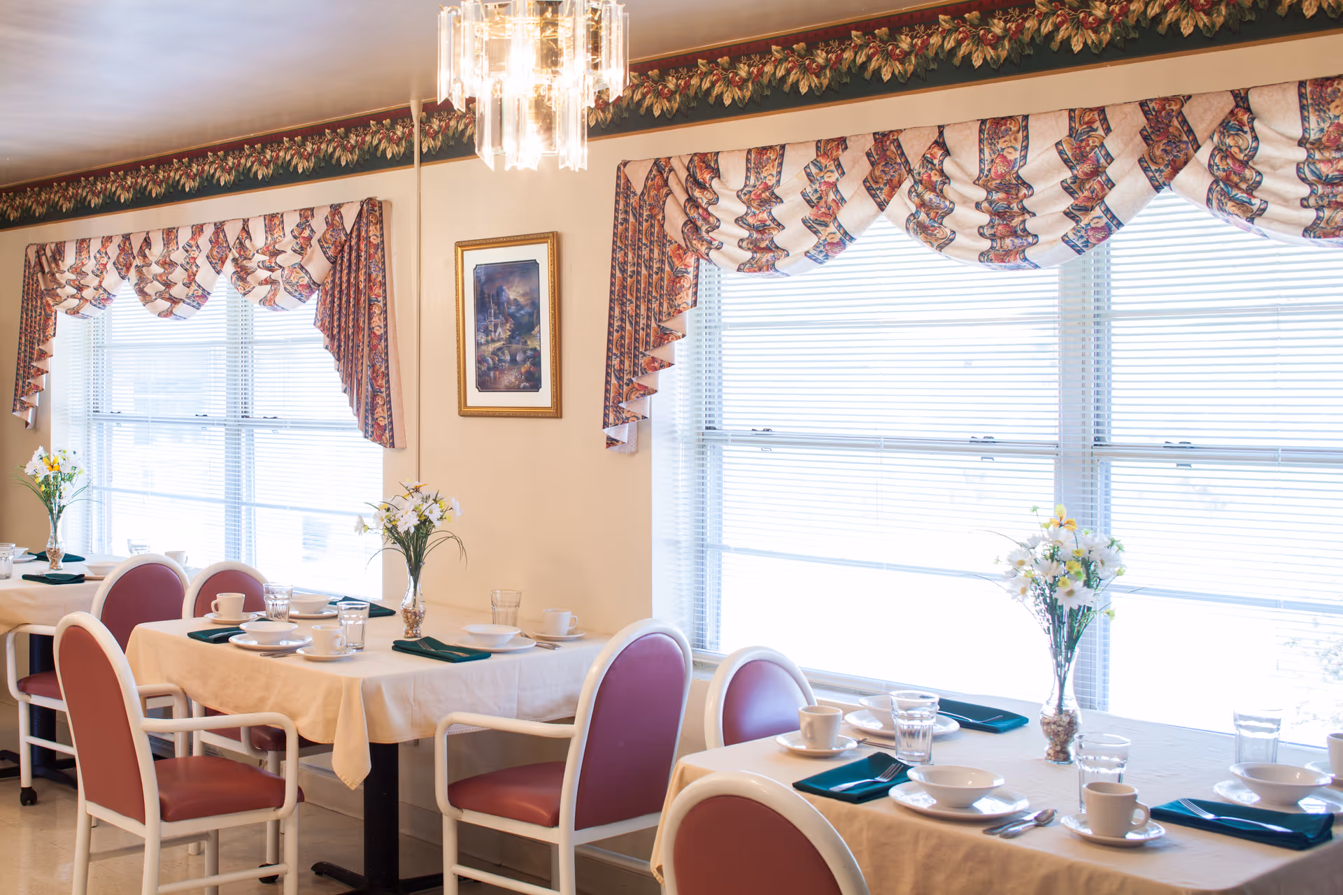 A bright dining room with tables set for a meal, featuring white tablecloths, pink cushioned chairs, green napkins, white plates, cups, and glassware. The room has large windows with floral valances and blinds, floral wallpaper border near the ceiling, a chandelier, and framed artwork on the wall.