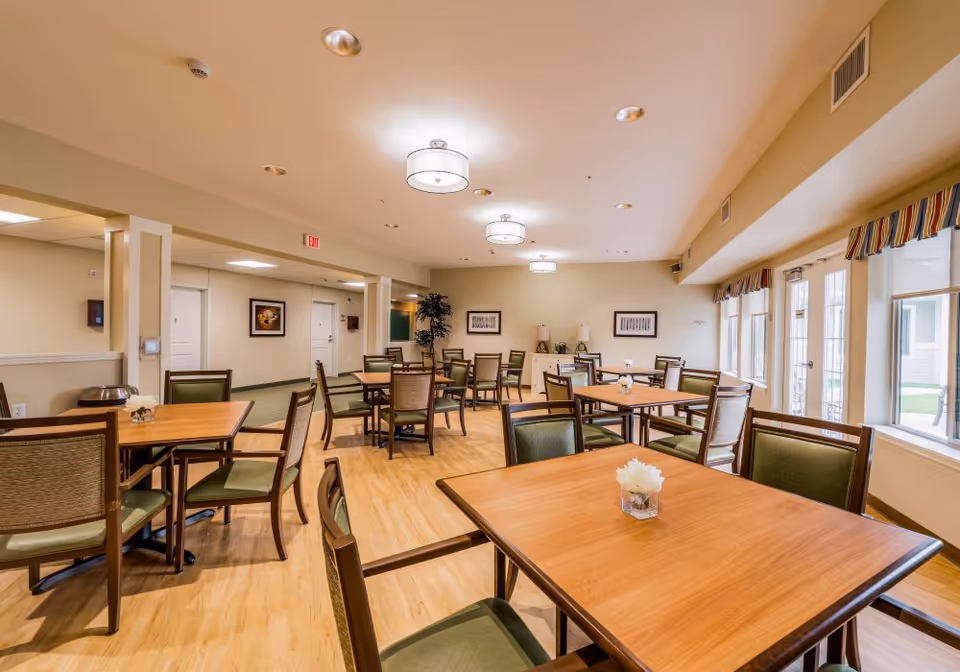 A bright and spacious dining room in a senior living facility with multiple wooden tables and green cushioned chairs arranged neatly. The room has light-colored walls, wooden flooring, ceiling lights, framed artwork on the walls, and large windows with striped valances letting in natural light.