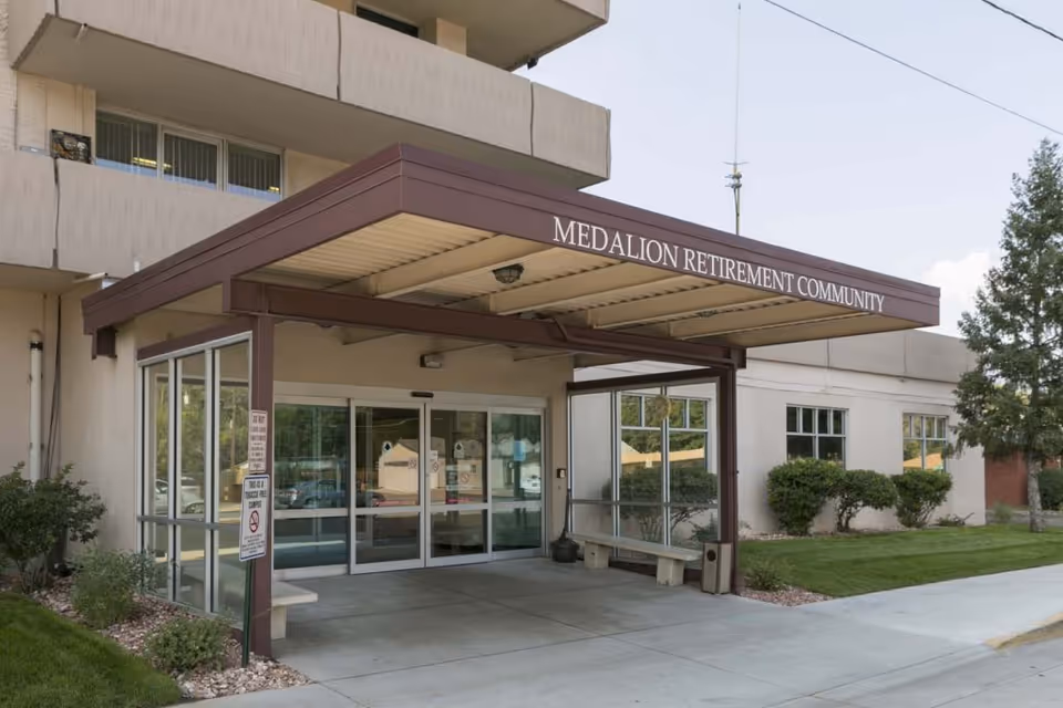 Entrance to Medallion Retirement Community with a covered walkway and automatic sliding glass doors, surrounded by bushes and a tree on a clear day.