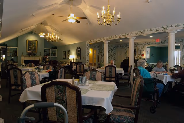 Dining room with set tables and upholstered chairs, chandeliers overhead, and several residents seated at tables.
