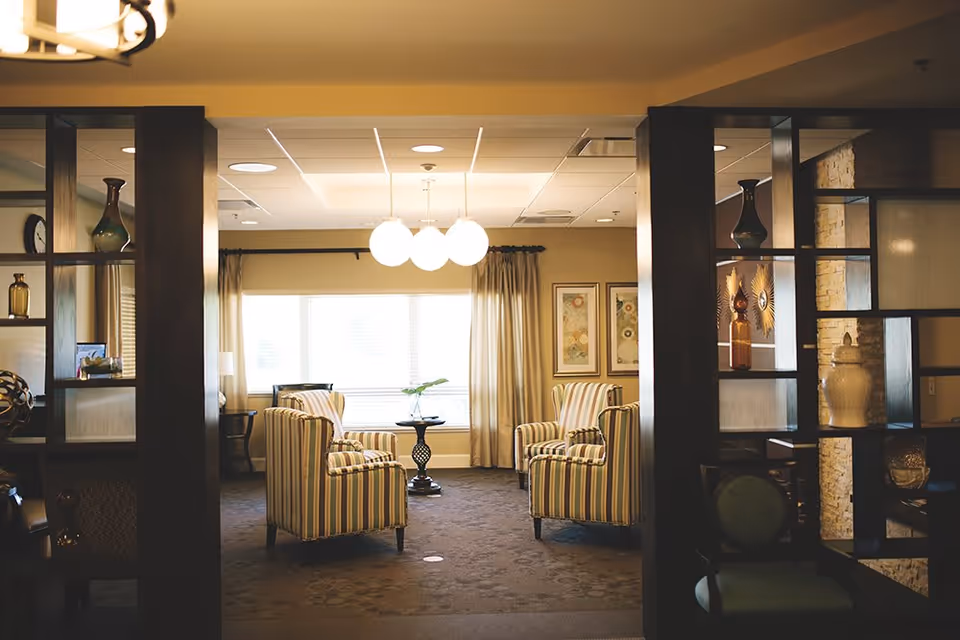 A cozy living room area with four striped armchairs arranged around a small round table with a plant on it. The room has large windows with curtains, two framed artworks on the wall, and a ceiling light fixture with three round bulbs. Dark wooden shelving units with decorative items flank the entrance to the room.