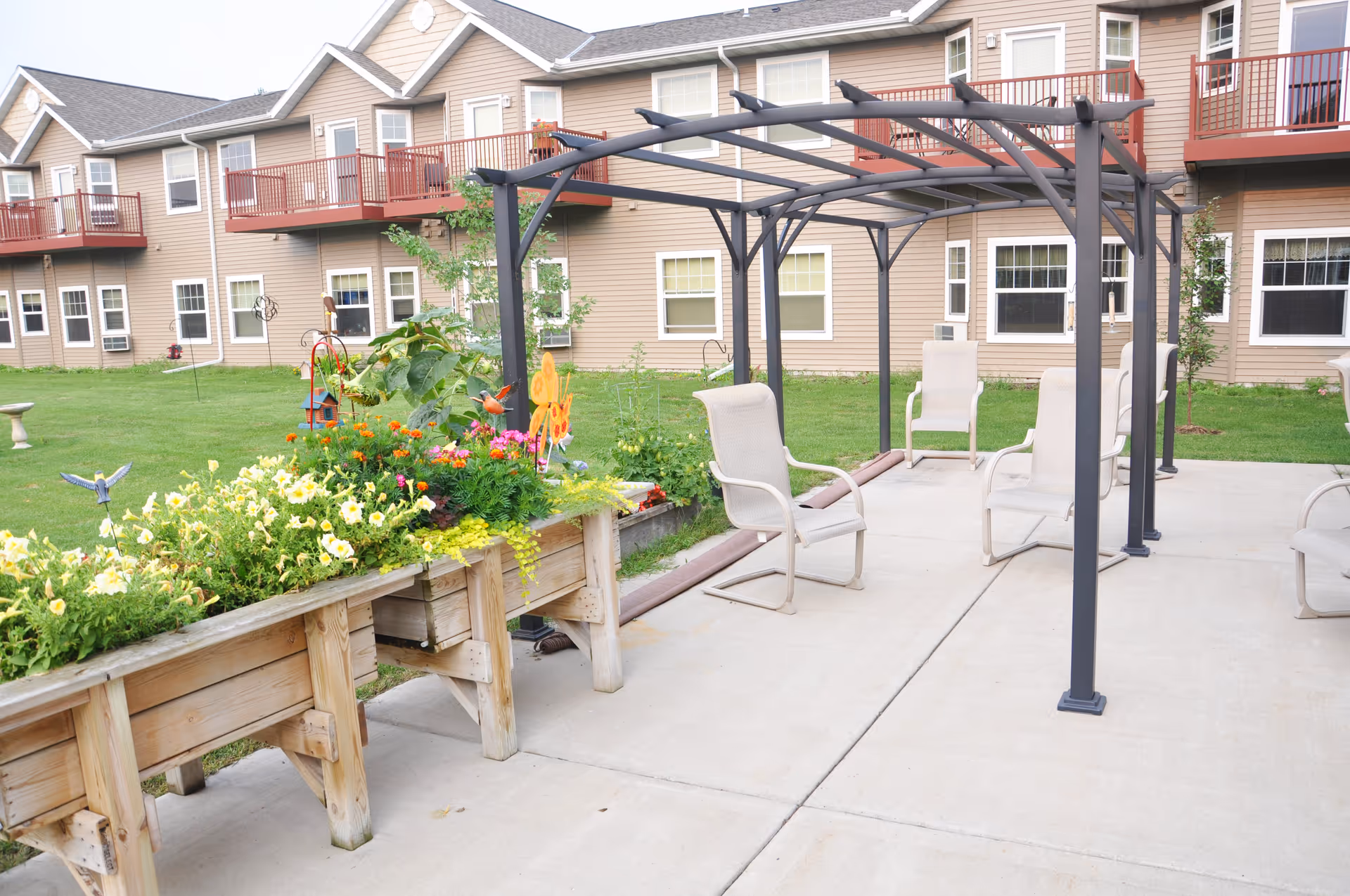 Outdoor courtyard with a pergola, patio chairs, raised wooden planters filled with flowers, and the apartment-style building in the background.