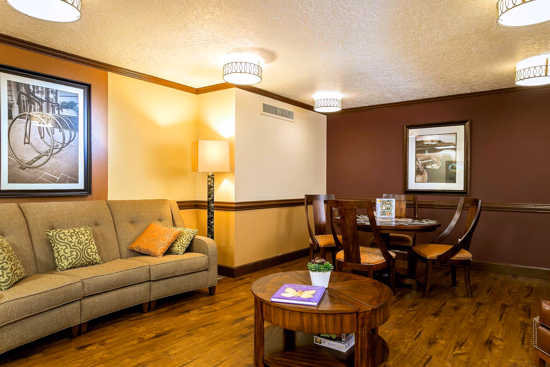 A cozy living room area with a brown sofa adorned with patterned cushions, a wooden round coffee table with a small potted plant and a book on top, and a dining table with four wooden chairs with orange cushions. The walls are painted in warm tones of beige and brown, decorated with framed black and white photographs. The room has wooden flooring and ceiling lights with decorative covers.