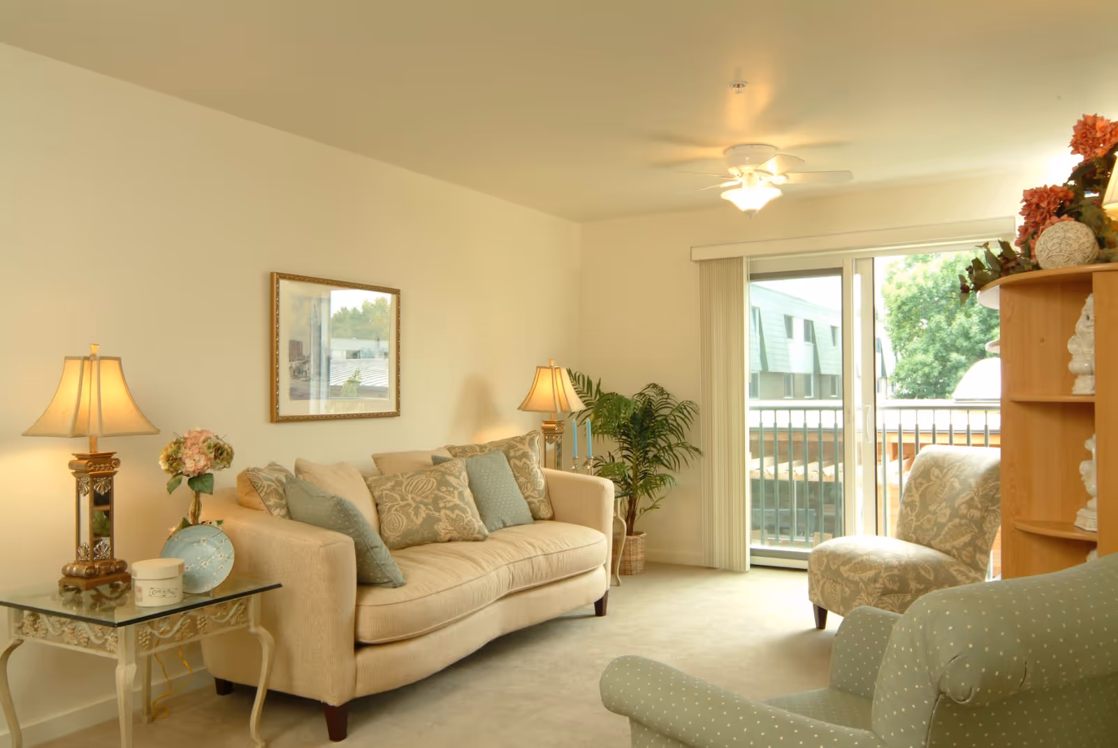 A cozy living room with a beige sofa adorned with patterned cushions, a glass side table with a lamp and decorative items, two upholstered chairs, a wooden shelving unit with floral decorations, a ceiling fan with light, and a sliding glass door leading to a balcony with a view of trees and neighboring buildings.