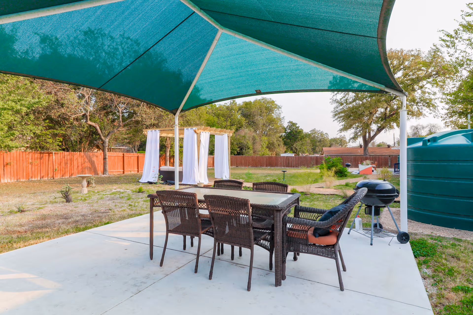 Outdoor patio area with a glass-top table and six wicker chairs under a large green canopy. In the background, there is a wooden pergola with white curtains, a charcoal grill, a large green water tank, and a fenced yard with trees and grass.