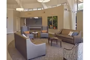 A spacious and well-lit living room area with modern furniture including two sofas, two armchairs, a coffee table, and side tables. The walls are light-colored with two framed pictures and a large window. The room has a carpeted floor and overhead lighting fixtures.