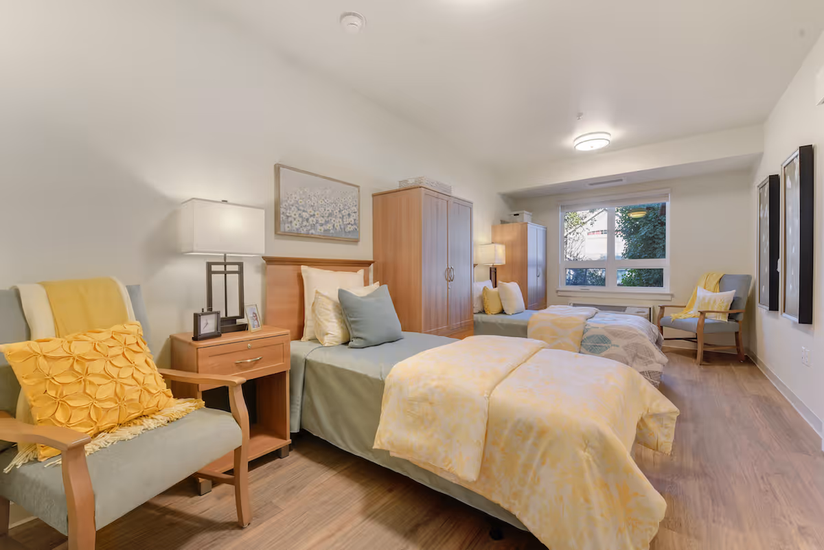 A bright, neatly arranged bedroom in a senior living facility with two single beds, each with light yellow and gray bedding. There are wooden nightstands with lamps and picture frames between the beds. Two armchairs with yellow cushions are placed near the beds. A large window lets in natural light, and the room has wooden flooring and light-colored walls with framed artwork.