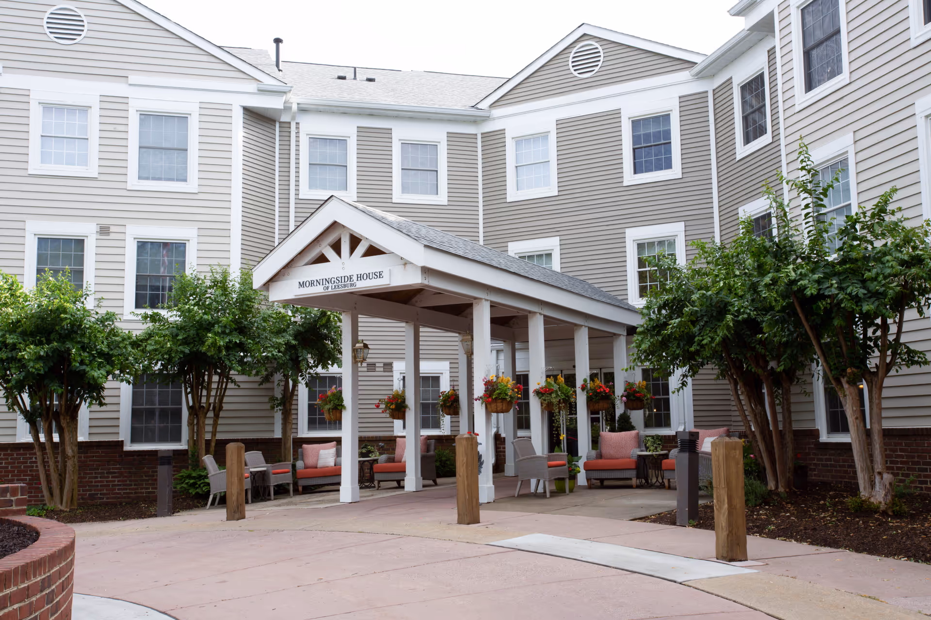 Entrance area of Morningside House of Leesburg featuring a covered porch with hanging flower baskets, outdoor seating with cushioned chairs and sofas, surrounded by a beige multi-story building with white trim and several windows, and landscaped with small trees and shrubs.