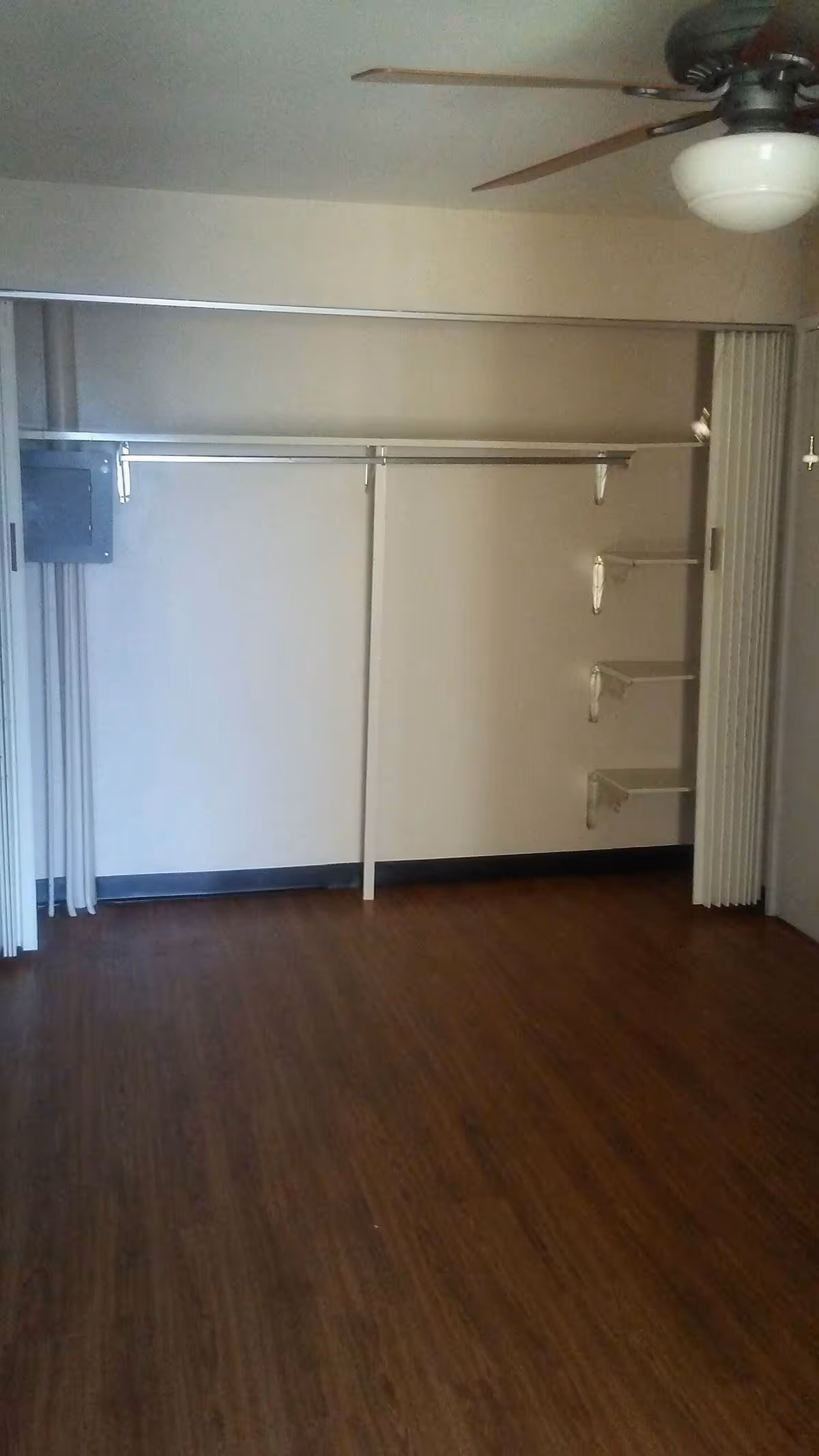 Empty room with wooden floor, a ceiling fan with light, and an open closet with a hanging rod and three small shelves on the right wall.