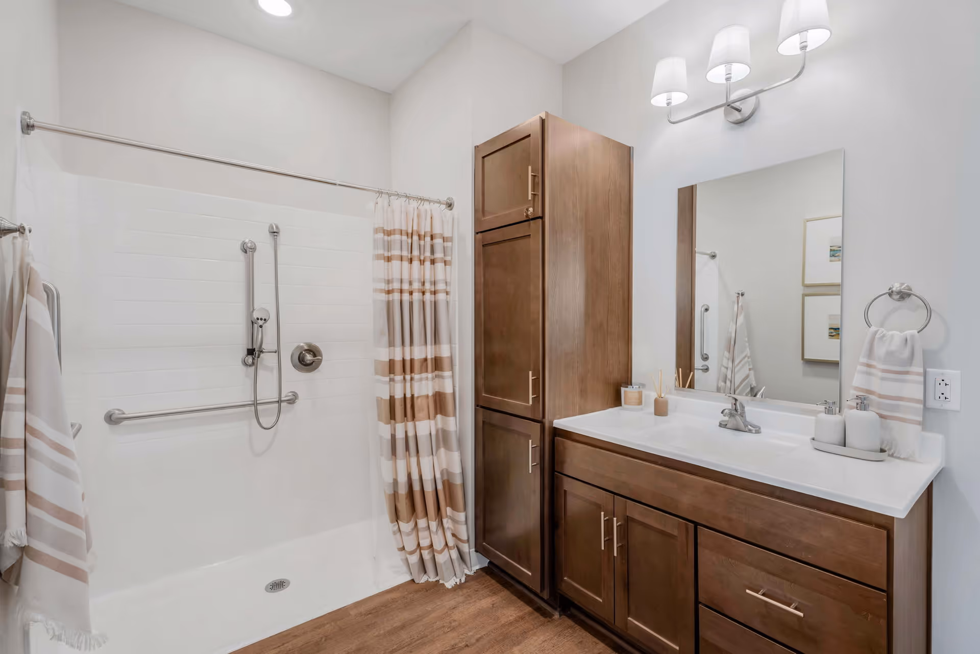 A modern bathroom with a walk-in shower featuring a handheld showerhead and grab bars. The shower has a striped beige and white curtain. Next to the shower is a wooden cabinet and a vanity with a white countertop, a sink, and a large mirror above. The vanity is decorated with a soap dispenser, a small container, and a reed diffuser. The walls are painted white and the floor has a wood-like finish.