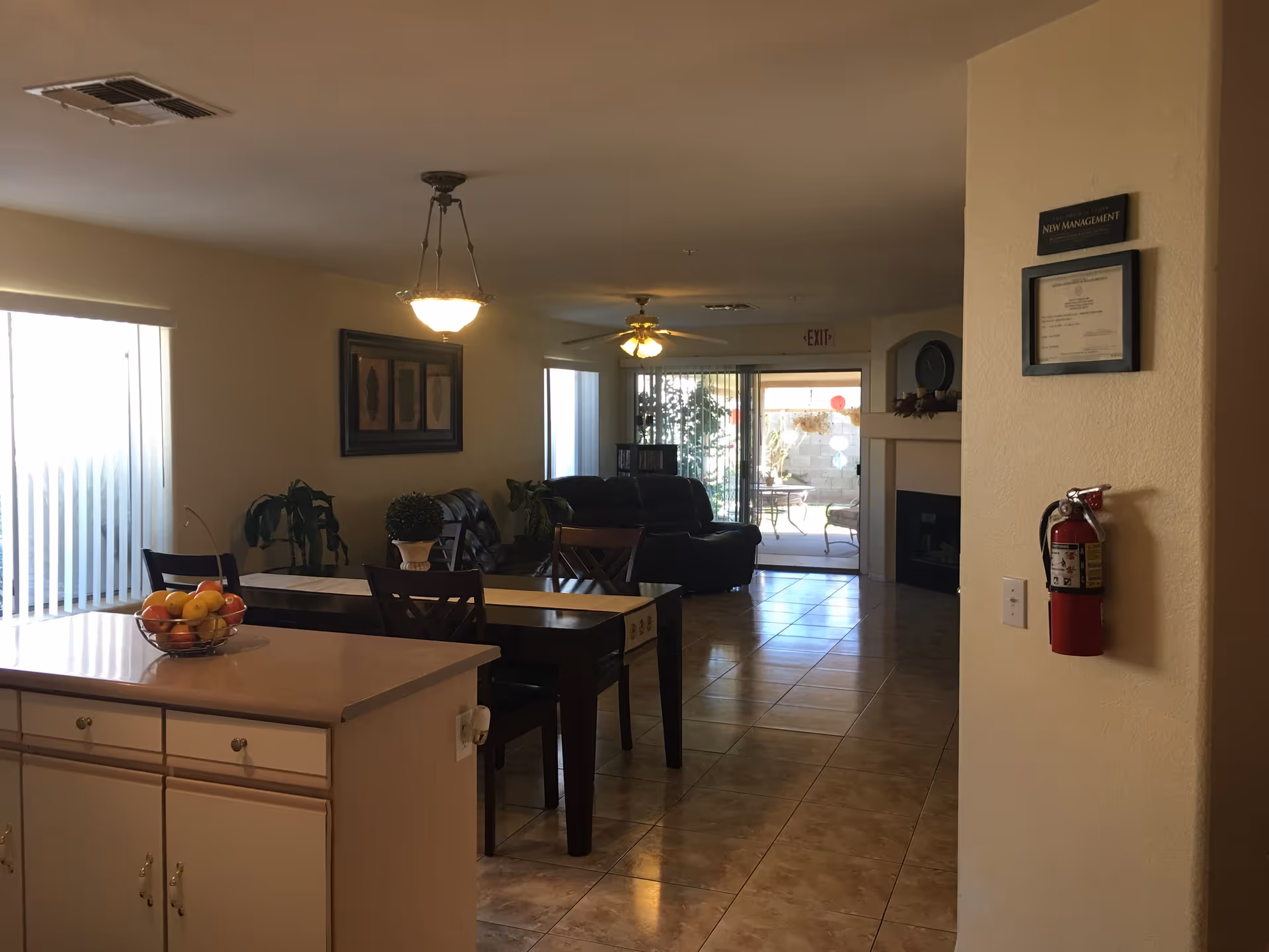 Interior view of a senior living facility showing a dining area with a table and chairs, a kitchen counter with a bowl of fruit, a living room with leather sofas, ceiling lights, a ceiling fan, a fireplace, and a sliding glass door leading to an outdoor patio.