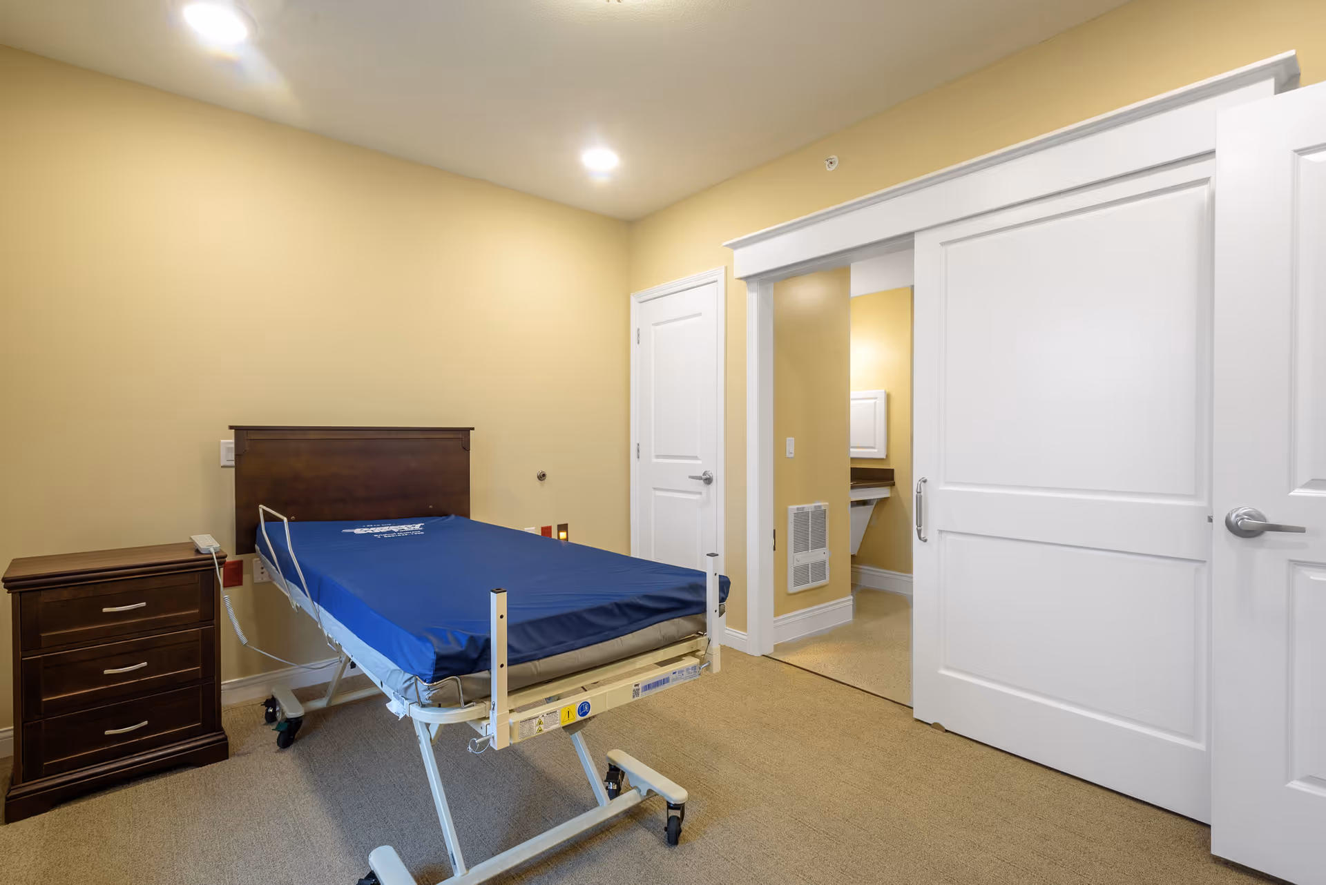 A simple room with a hospital-style bed with a blue mattress, a wooden nightstand with three drawers, beige walls, and carpeted floor. There are two white doors, one sliding door partially open revealing a sink area, and another door closed.