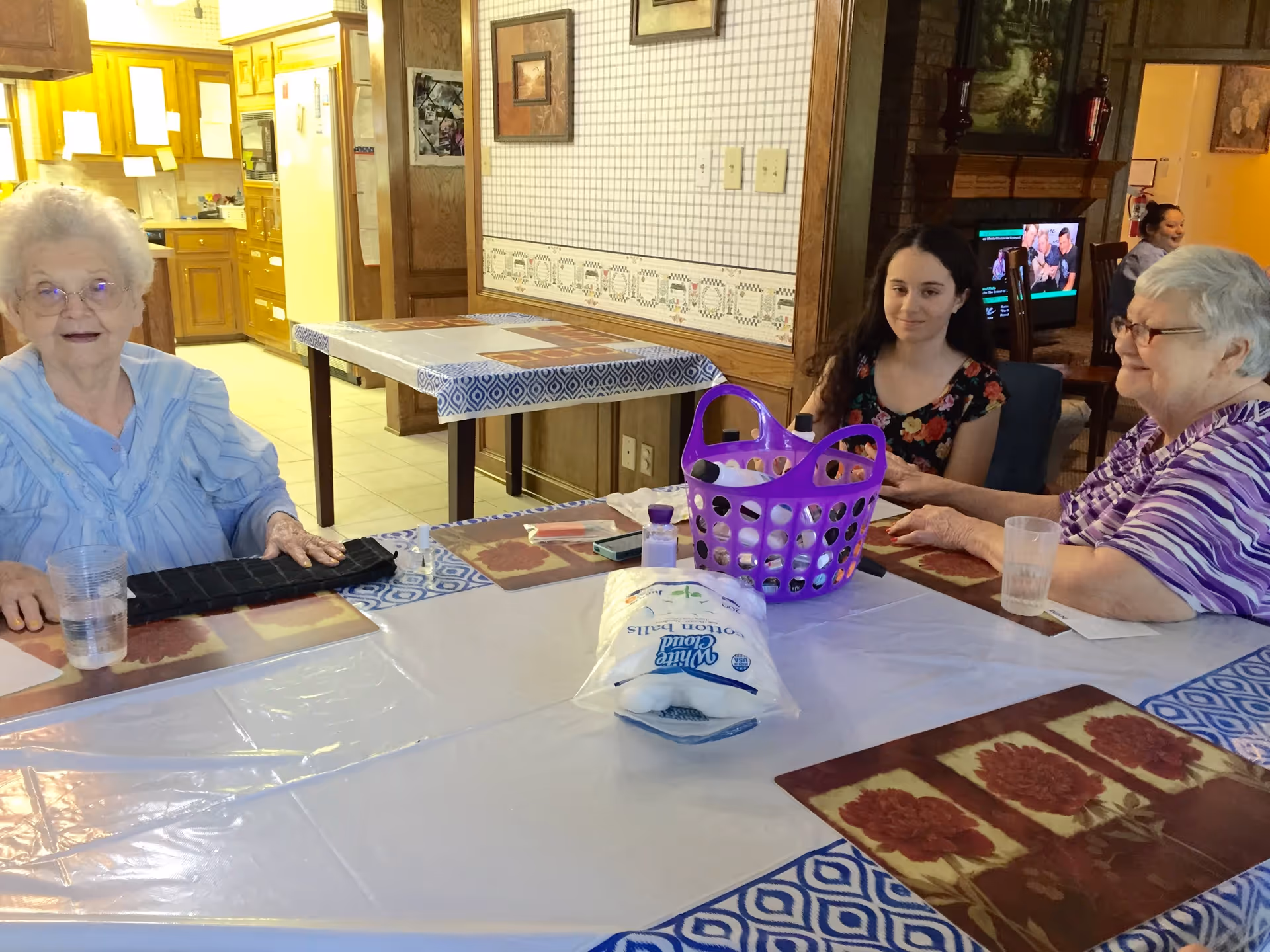 Three women sitting around a dining table in a room with a kitchen in the background. The table is covered with a plastic tablecloth and has placemats, a purple basket, a bag of cotton balls, and some small bottles on it. The room has wooden cabinets and wallpaper with a floral border.
