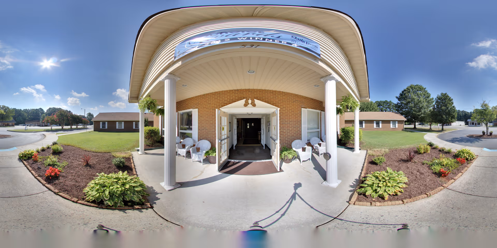 Front entrance of a brick senior living building with a covered portico, white columns, chairs, and landscaped flower beds.