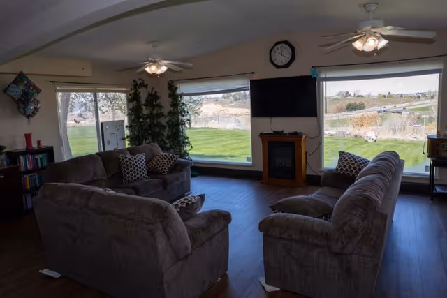 A cozy living room with three gray upholstered sofas arranged around a small wooden electric fireplace and a wall-mounted flat-screen TV. The room has large windows offering a view of a green lawn and a distant landscape. Ceiling fans with lights are mounted on the ceiling, and decorative pillows are placed on the sofas.
